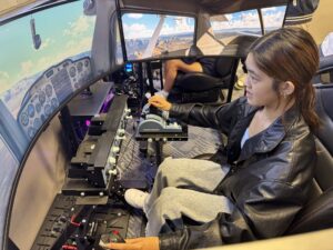 A young person operating flight simulator controls, with aircraft instruments and a virtual landscape displayed across multiple screens.